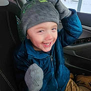 Aiden is registered to the contest to win money with this photo: child, smiling, winter_clothing, hat, mittens, blue_jacket, car_interior, seat, window, snow, outdoor, happy, young_child, warm_clothing, green_pom_pom, casual, portrait, seated, joyful, playful