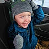 child, smiling, winter_clothing, hat, mittens, blue_jacket, car_interior, seat, window, snow, outdoor, happy, young_child, warm_clothing, green_pom_pom, casual, portrait, seated, joyful, playful