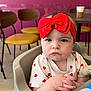 baby, child, headband, red_bow, high_chair, clothing, hands, face, indoor, wooden_chair, table, coffee_cup, expressionless, person, infant, casual_clothing, flooring, pink_wall, furniture, portrait