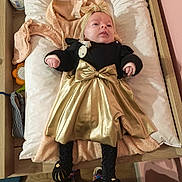 Emy a rejoint le concours — aidez-le/la à gagner de superbes lots ! baby, infant, clothing, gold_skirt, headband, bow, blanket, changing_pad, black_top, leggings, knitted_booties, wooden_surface, floor, person, indoor, cute, lying_down, soft_toys, towel, baby_care