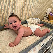 Isabella is registered to the contest to win money with this photo: baby, smiling, diaper, changing_table, wallpaper, floral_pattern, indoor, infant, happy, cute, skin, furniture, room, blanket, person, head, face, child, lying_down, mohawk