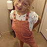 child, girl, glasses, smiling, posing, indoor, carpet, mirror, white_shirt, pink_overalls, sandals, blonde_hair, happy, person, fashion, footwear, wall, door, room, casual
