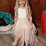 child, blonde_hair, dress, barefoot, indoor, wooden_furniture, floor, smile, long_hair, girl, toy, colorful, gift_bag, curly_hair, person, portrait, happy, young, standing, home