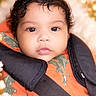 baby, infant, child, car_seat, dinosaur_print, orange_clothing, curly_hair, face, portrait, close_up, cute, seatbelt, soft_lighting, bokeh, indoors, young_child, expressionless, person, baby_boy, warm_colors