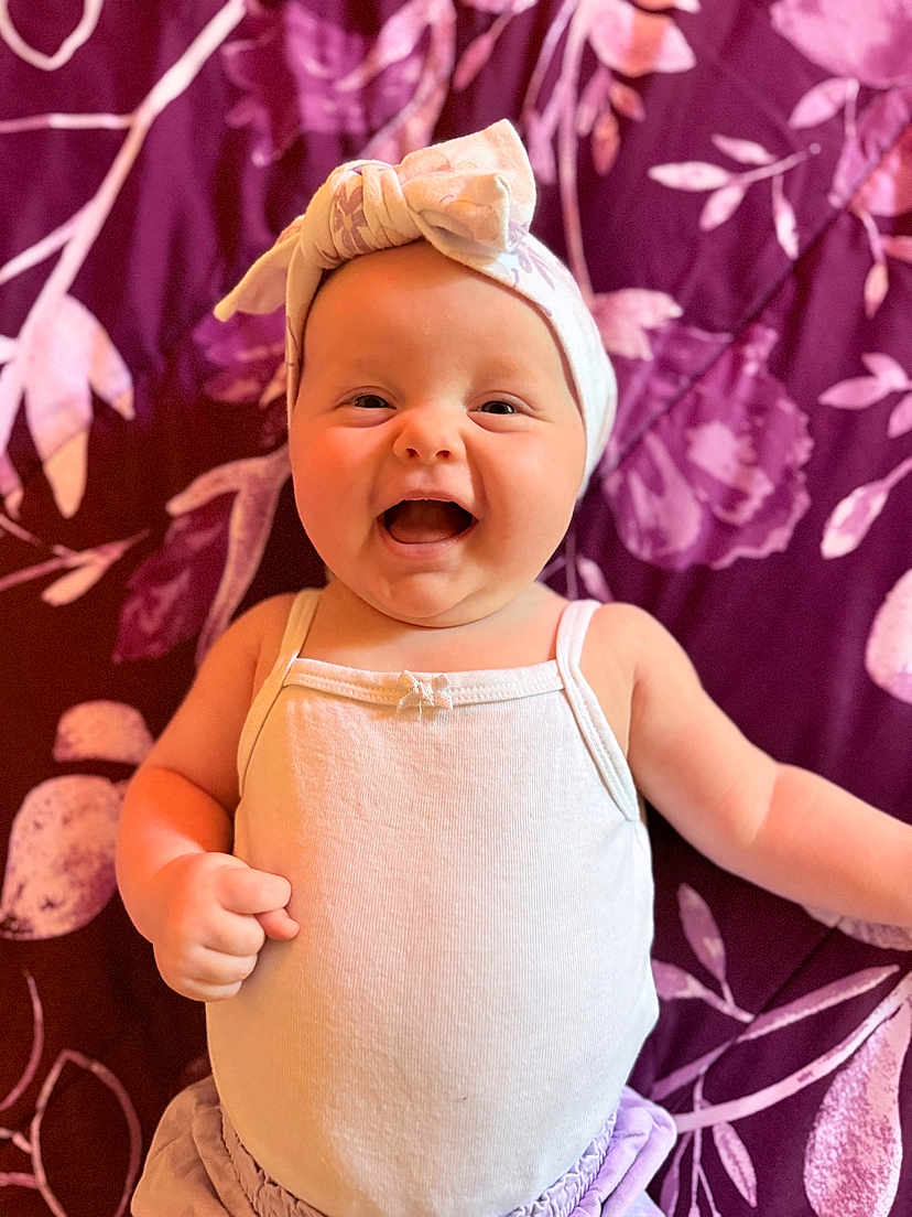 Kaya is registered to the contest to win money with this photo: baby, infant, smiling, happy, headwrap, white_clothing, purple_background, flower_pattern, cute, portrait, child, skin, face, arm, hand, clothing, indoors, young, person, expression