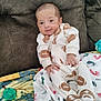 baby, infant, onesie, football_pattern, blanket, couch, indoor, child, footwear, fabric, pattern, relaxed, cute, small_hands, soft_texture, seated, comfort, expression, face, head