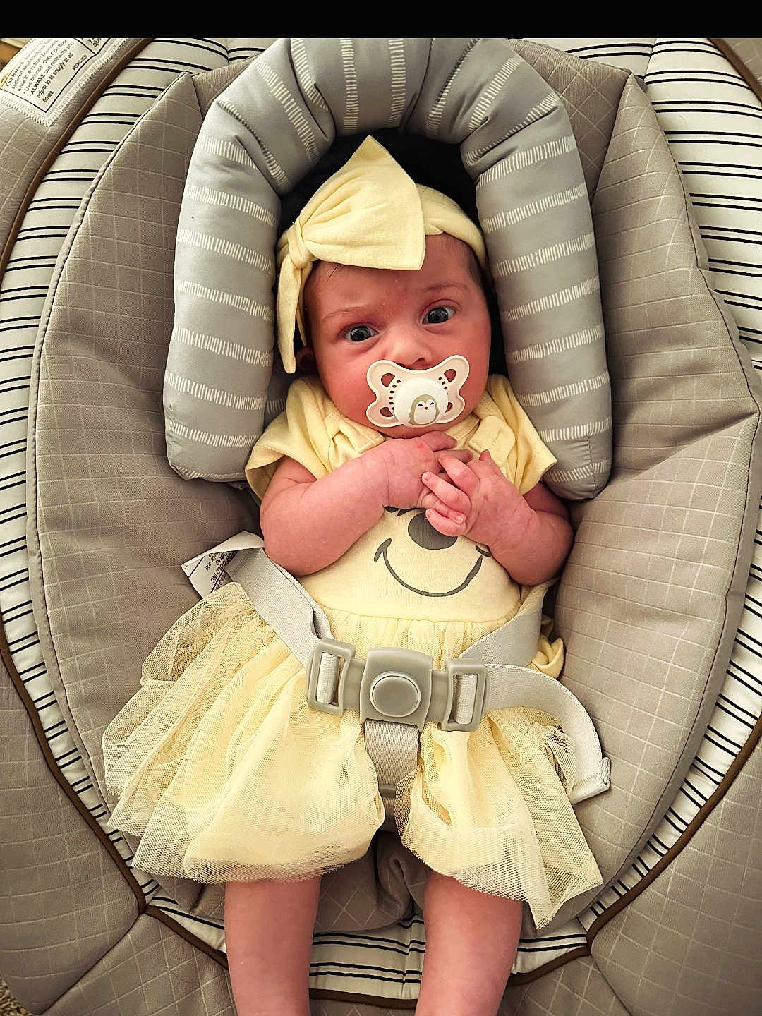 Ellie is registered to the contest to win money with this photo: baby, infant, pacifier, yellow_dress, headband, baby_seat, seatbelt, cushion, striped_pattern, smile_print, cute, small_hands, wide_eyes, indoors, child, newborn, comfortable, soft_fabric, adorable, tulle_skirt