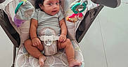 Hanah is registered to the contest to win money with this photo: baby, infant, child, bouncer, baby_seat, buckled, sitting, wide_eyed, curly_hair, gray_shirt, toys, toy_bar, indoor, tiled_floor, playroom, portrait, full_body, chair, sandal, photography