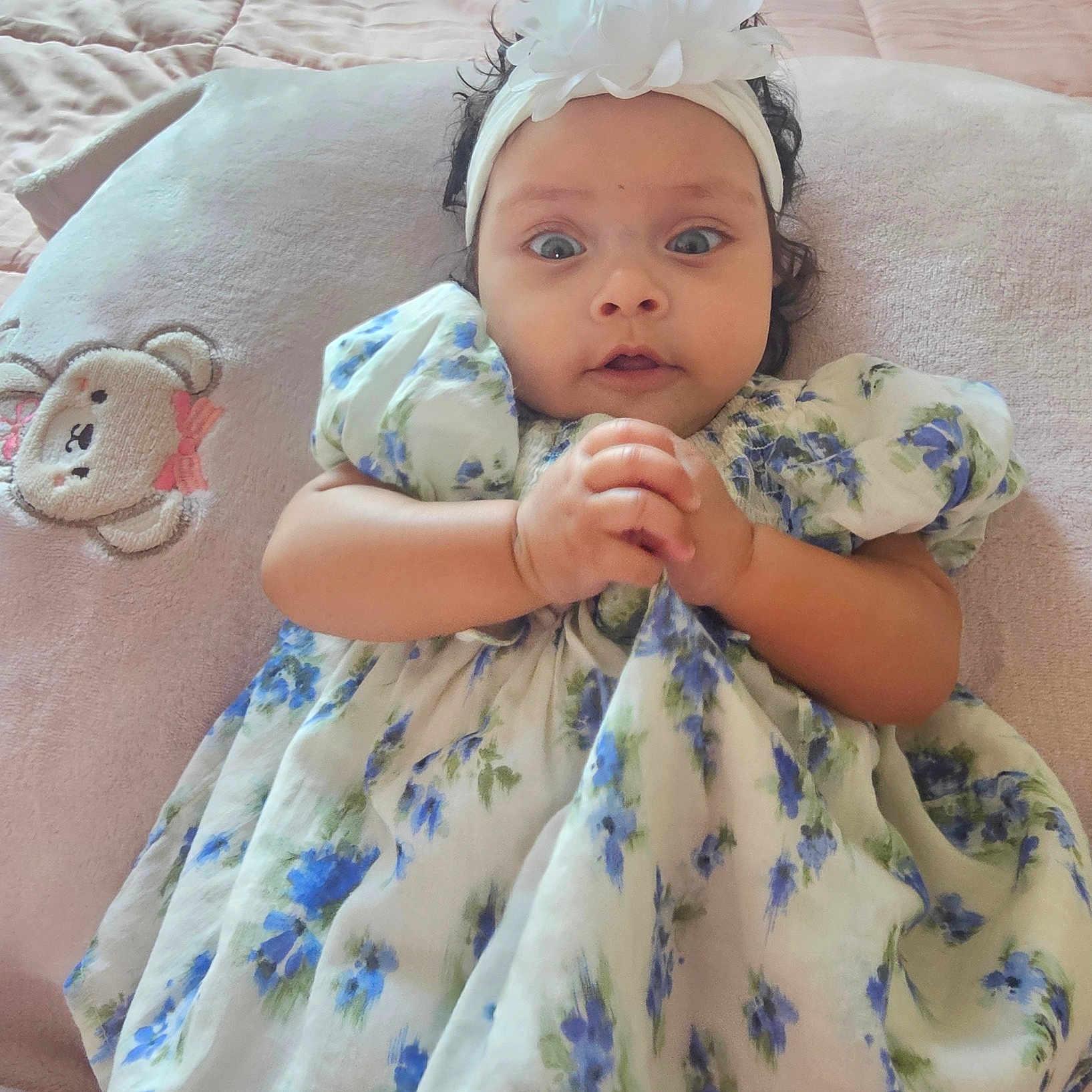 Hanah joined the competition — help win amazing prizes! baby, child, infant, portrait, wide_eyed, floral_dress, headband, flower, teddy_bear, pillow, bed, indoors, lying_down, hands_clasped, curly_hair, smiling, cute, soft_lighting, baby_fashion, close_up
