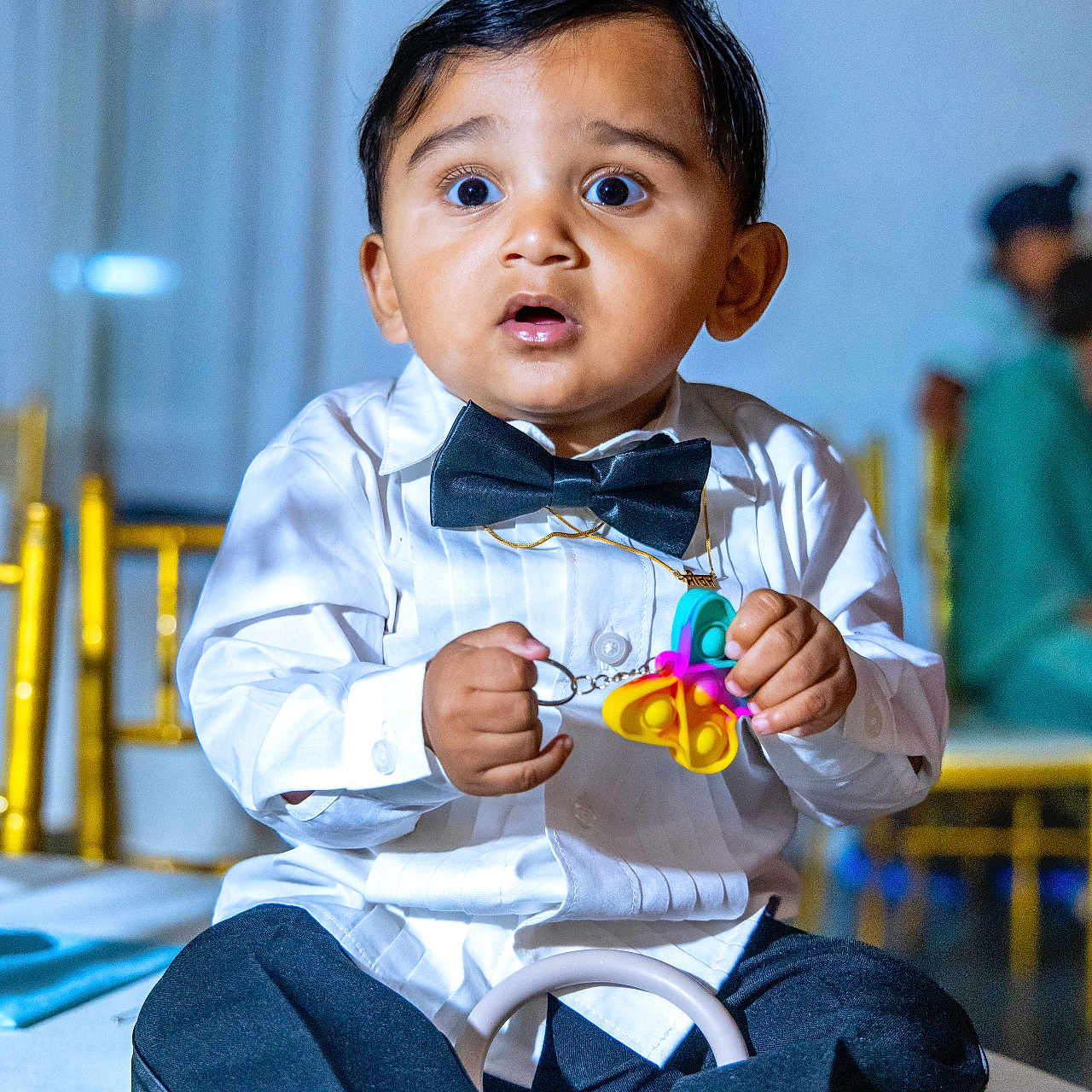 Siraj is registered to the contest to win money with this photo: black_pants, blue_stripes, bow_tie, boy, butterfly_toy, child, curious, cute, event, expression, formal_wear, indoor, people_in_background, portrait, sitting, socks, toddler, toy, white_shirt, wide_eyes