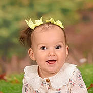 Diora is registered to the contest to win money with this photo: baby, child, portrait, smiling, happy, face, eyes, mouth, hair_bows, yellow_bows, ponytail, floral_dress, collar, clothing, sitting, front_facing, outdoor_background, blurred_background, studio_portrait, infant