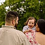 back_view, beard, casual_clothing, child, curly_hair, family, floral_dress, greenery, happy, holding, love, man, nature, outdoor, parent, portrait, smiling, toddler, together, woman
