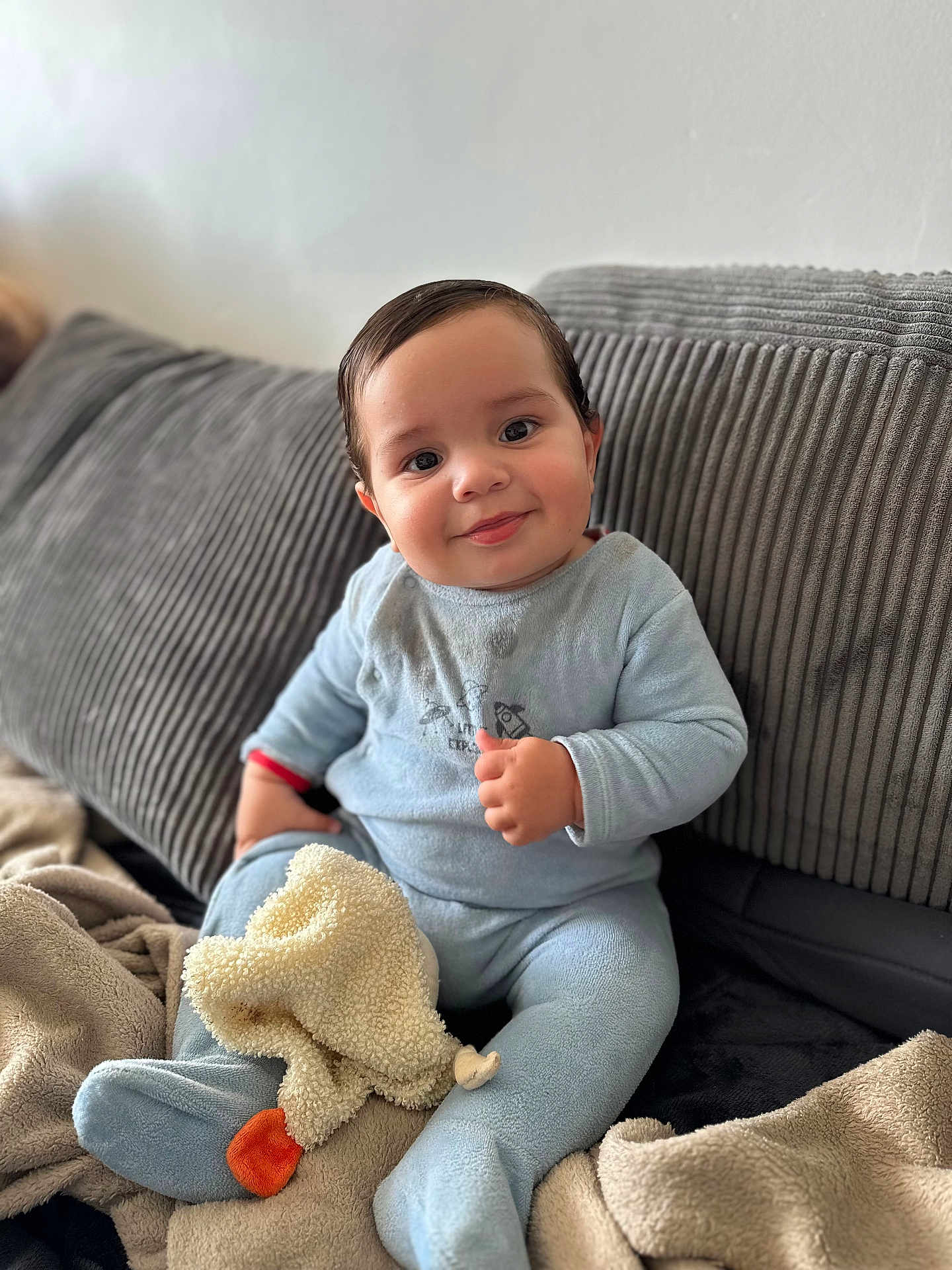 Ezio a rejoint le concours — aidez-le/la à gagner de superbes lots ! baby, child, smile, thumbs_up, blue_onesie, blanket, couch, soft_toys, indoor, cute, happy, face, person, seated, plush, cozy, infant, clothing, home, portrait