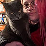 cat, person, portrait, selfie, indoors, black_cat, gray_fur, pink_hair, glasses, nose_ring, cuddling, pet, animal, whiskers, yellow_eyes, sitting, close_up, home, cabinet, casual