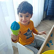 Anis a rejoint le concours — aidez-le/la à gagner de superbes lots ! child, smiling, indoor, play_dough, green_cone, yellow_shirt, gray_pants, curtains, table, wood_floor, chair, face, happy, messy, toy, playtime, casual_clothing, young_child, person, daylight