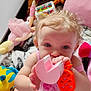 baby, child, toys, doll, stuffed_animal, pink_clothing, teether, curly_hair, blue_eyes, indoor, play_area, blanket, soft_toys, cute, infant, sitting, colorful, closeup, playtime, fun