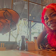 Ezekiel is registered to the contest to win money with this photo: child, woman, pink_hair, glasses, smile, selfie, restaurant, table, condiments, bottle, window, napkin, interior, natural_light, family_moment, portrait, casual, happy, dining, decor
