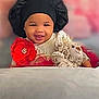 baby, child, smiling, black_bonnet, red_flower, plush_toy, teddy_bear, pillow, red_roses, soft_toys, indoors, cute, happy, portrait, infant, clothing, face, headwear, decor, cozy