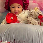 Heavenly is registered to the contest to win money with this photo: baby, child, smiling, bonnet, flower, red_flower, pillow, teddy_bear, heart, crown, plush_toy, indoor, cute, portrait, infant, soft_toy, decor, happy, headwear, lying_down