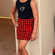 Isreãl is registered to the contest to win money with this photo: child, girl, indoor, posing, black_top, heart_design, red_skirt, checkered_skirt, socks, grey_socks, carpet, wall, corner, standing, fashion, smiling, young, person, casual, home