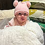 baby, pink_hat, pom_poms, blanket, pillow, floral_pattern, infant, cute, cozy, indoor, child, soft_texture, knitwear, smiling, face, portrait, toddler, warm_clothing, home, resting