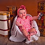baby, child, smiling, pink_headband, gingham_clothing, blanket, chair, wooden_floor, vintage_suitcases, brown_background, happy, sitting, portrait, indoor, cute, small_object, person, footwearless, head, infant