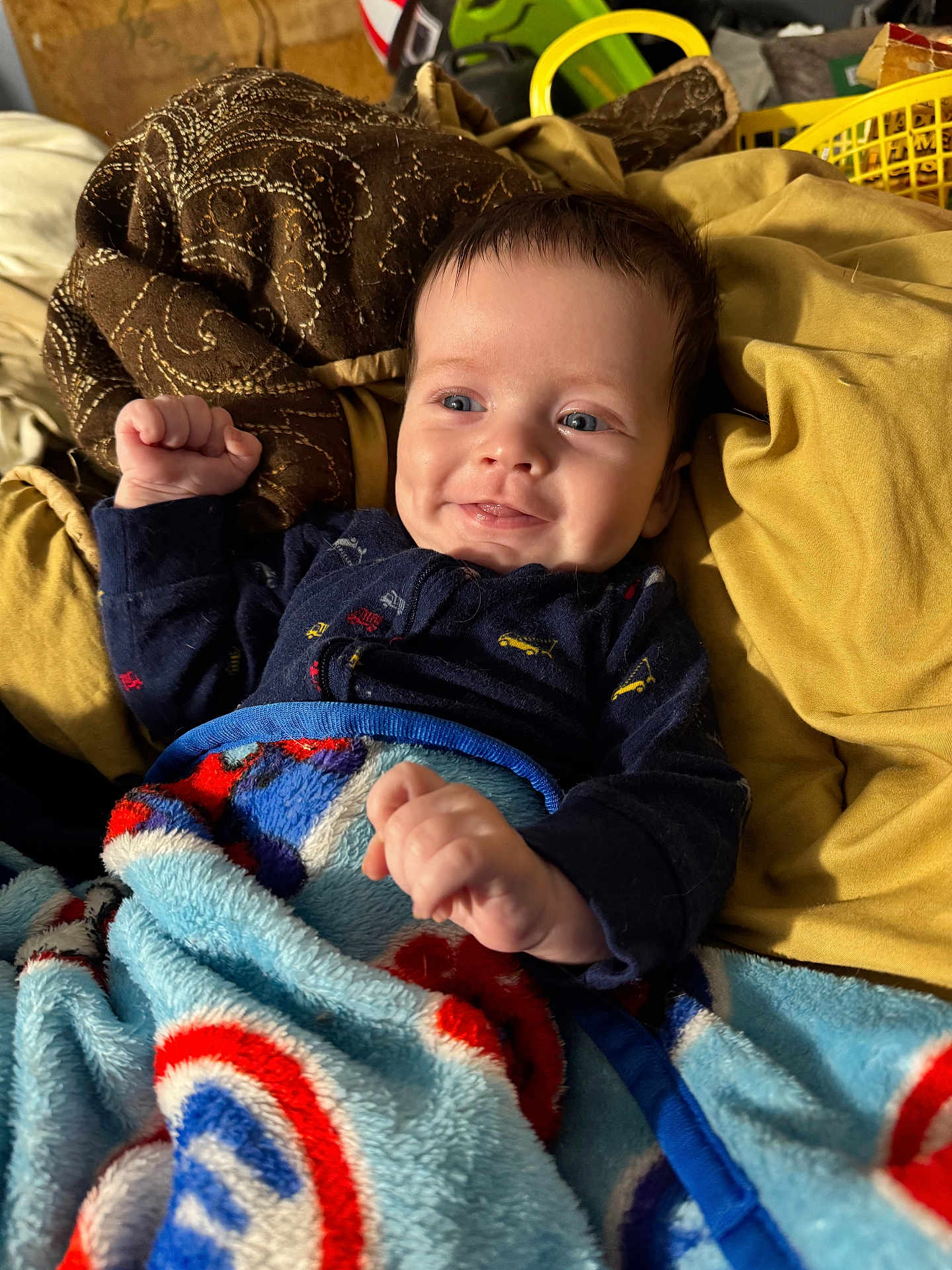 Denver is registered to the contest to win money with this photo: baby, smiling, blanket, blue, red, yellow, brown, clothing, infant, face, hands, pillow, comfortable, indoor, cute, portrait, happy, child, cozy, resting