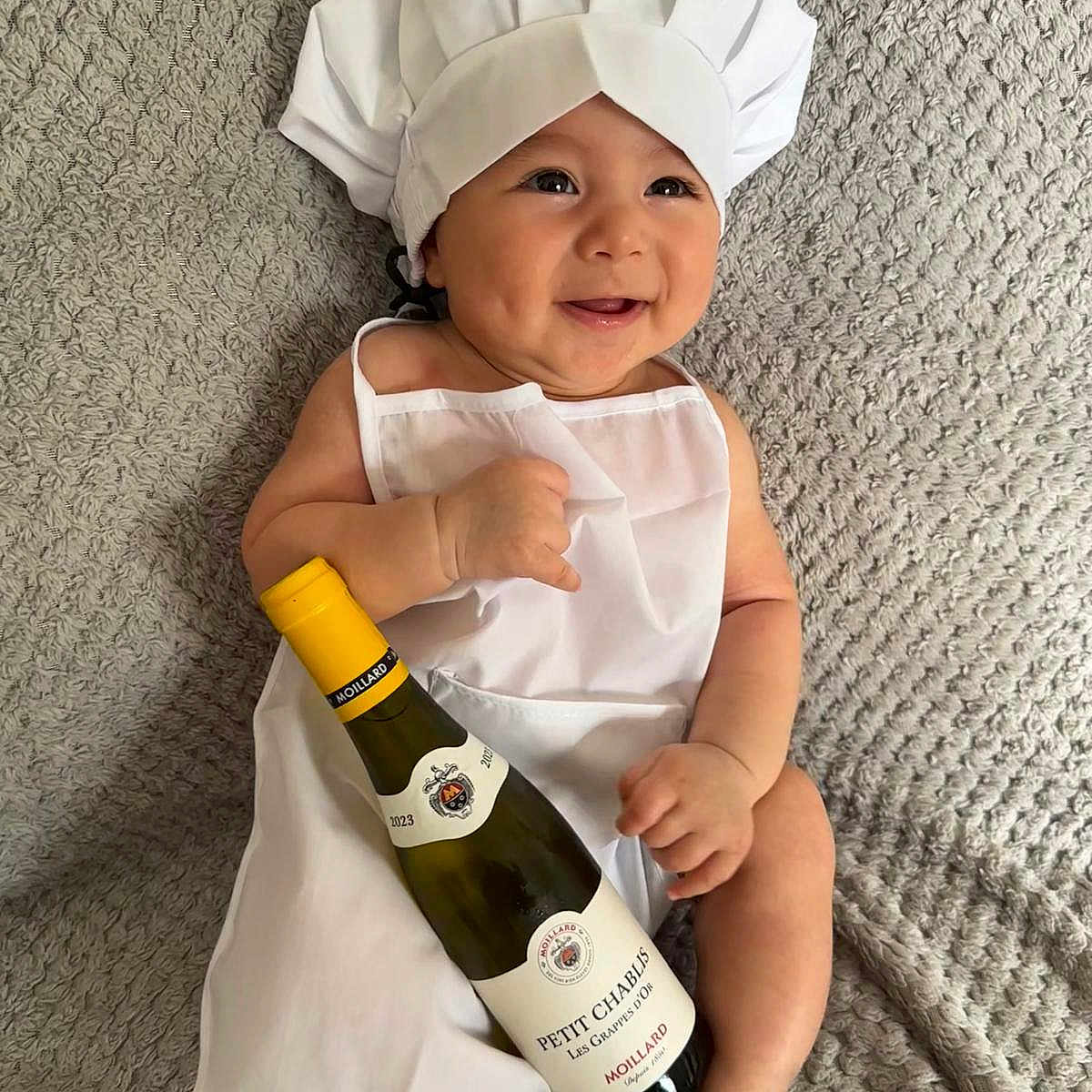 Hady joined the competition — help win amazing prizes! apron, baby, blanket, chef_hat, child, closeup, cute, happy, holding_object, indoors, infant, lying_down, person, playful, portrait, smiling, soft_lighting, texture, white_clothing, wine_bottle
