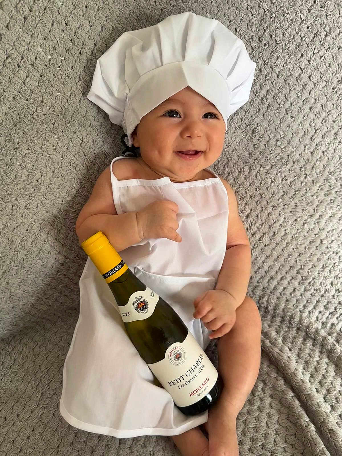 Hady joined the competition — help win amazing prizes! baby, chef_hat, apron, wine_bottle, smiling, infant, white_clothing, texture, blanket, cute, portrait, lying_down, holding_object, indoors, playful, happy, person, child, soft_lighting, closeup