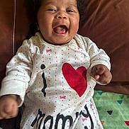 Makayla joined the competition — help win amazing prizes! baby, child, smiling, laughing, curly_hair, onesie, clothing, text, heart, love, couch, blanket, indoor, happy, infant, person, face, cute, joy, portrait