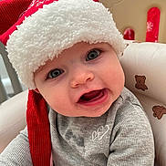 Ayla joined the competition — help win amazing prizes! baby, child, santa_hat, red_hat, gray_shirt, teddy_bear_pattern, chair, cute, face, expression, indoors, holiday, festive, smiling, portrait, person, infant, seat, cozy, playful