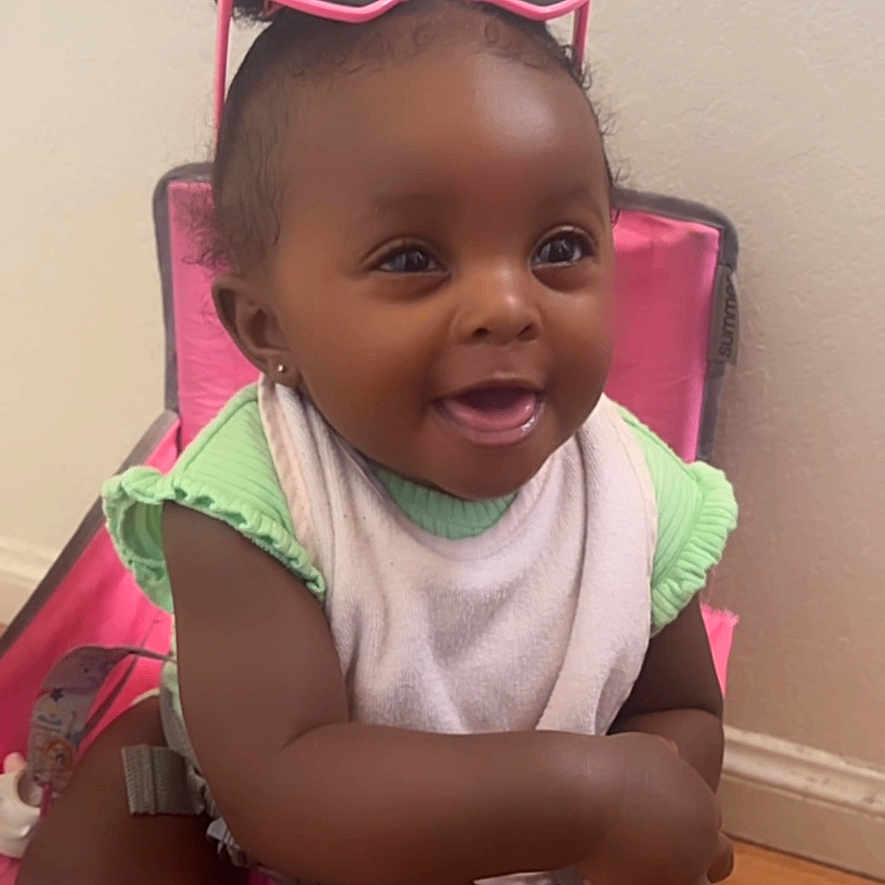 Jaziyah is registered to the contest to win money with this photo: accessory, baby, bib, casual, chair, child, curly_hair, cute, dark_skin, green_clothing, happy, indoor, infant, pink_sunglasses, playful, portrait, seated, smiling, socks, wooden_floor