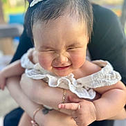 Mya joined the competition — help win amazing prizes! baby, child, infant, smiling, face, hair_accessory, white_clothing, cute, chubby_cheeks, person, adult_hands, holding, outdoor, blurred_background, portrait, happy, closeup, skin, expression, family