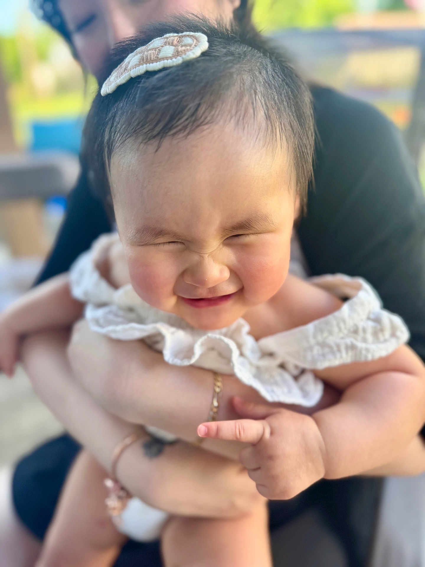 Mya joined the competition — help win amazing prizes! baby, child, infant, smiling, face, hair_accessory, white_clothing, cute, chubby_cheeks, person, adult_hands, holding, outdoor, blurred_background, portrait, happy, closeup, skin, expression, family