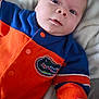 baby, infant, newborn, onesie, orange_clothing, blue_clothing, blanket, bedding, face, eyes, hand, portrait, close_up, cute, sleepy, gator_patch, sports_logo, fabric, indoor, soft_texture