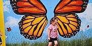 Emerie joined the competition — help win amazing prizes! child, girl, butterfly_wings, mural, outdoor, grass, gravel, sky, clouds, pink_water_bottle, shorts, tshirt, shoes, curly_hair, art, painting, nature, playful, colorful, wall
