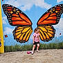 child, girl, butterfly_wings, mural, outdoor, grass, gravel, sky, clouds, pink_water_bottle, shorts, tshirt, shoes, curly_hair, art, painting, nature, playful, colorful, wall