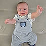 baby, smiling, striped_overalls, bear_patch, white_shirt, reaching_hands, lying_down, beige_background, cute, infant, happy, child, portrait, indoors, soft_surface, playful, face, head, person, clothing