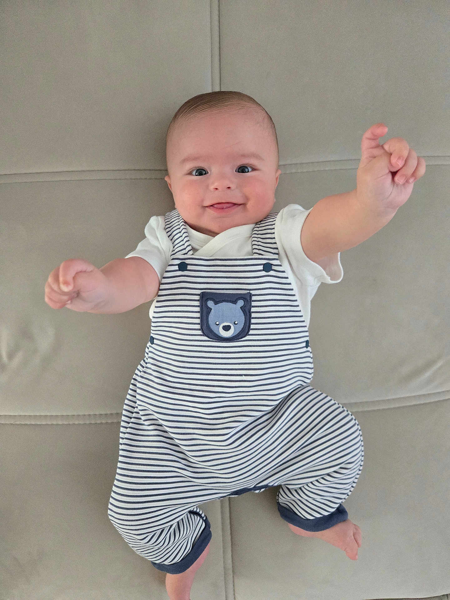 Jack joined the competition — help win amazing prizes! baby, smiling, striped_overalls, bear_patch, white_shirt, reaching_hands, lying_down, beige_background, cute, infant, happy, child, portrait, indoors, soft_surface, playful, face, head, person, clothing