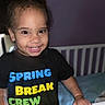toddler, child, smiling, face, hair_buns, black_tshirt, colorful_text, crib, bedroom, purple_wall, indoors, happy, cute, person, young_child, portrait, hand, clothing, furniture, baby