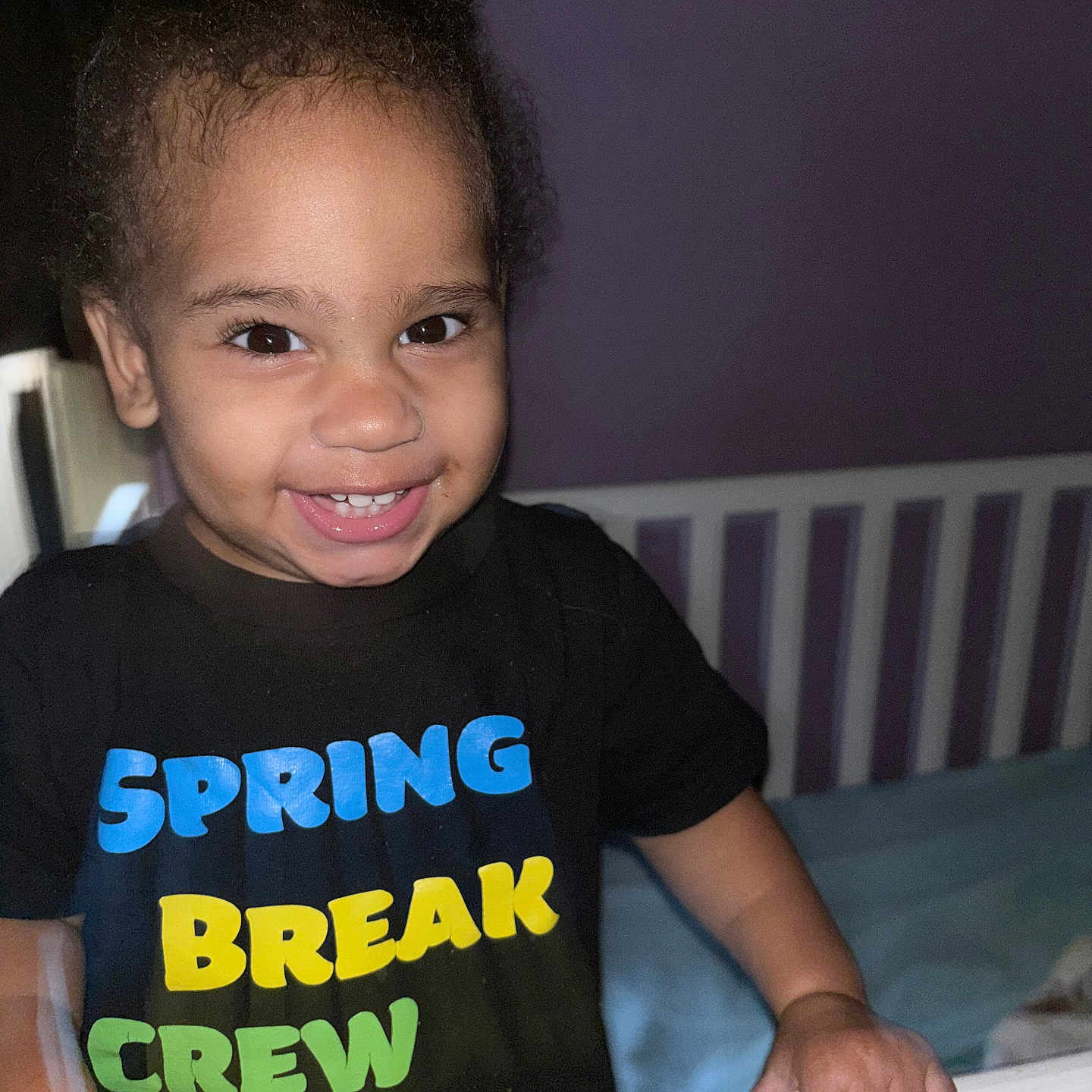 Nisaiah joined the competition — help win amazing prizes! toddler, child, smiling, face, hair_buns, black_tshirt, colorful_text, crib, bedroom, purple_wall, indoors, happy, cute, person, young_child, portrait, hand, clothing, furniture, baby