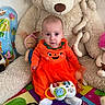 baby, child, orange_dress, pumpkin_outfit, toy, stuffed_animal, teddy_bear, play_mat, indoor, colorful, plush, sitting, cute, face, infant, soft_toys, bright_colors, young_child, expression, cozy