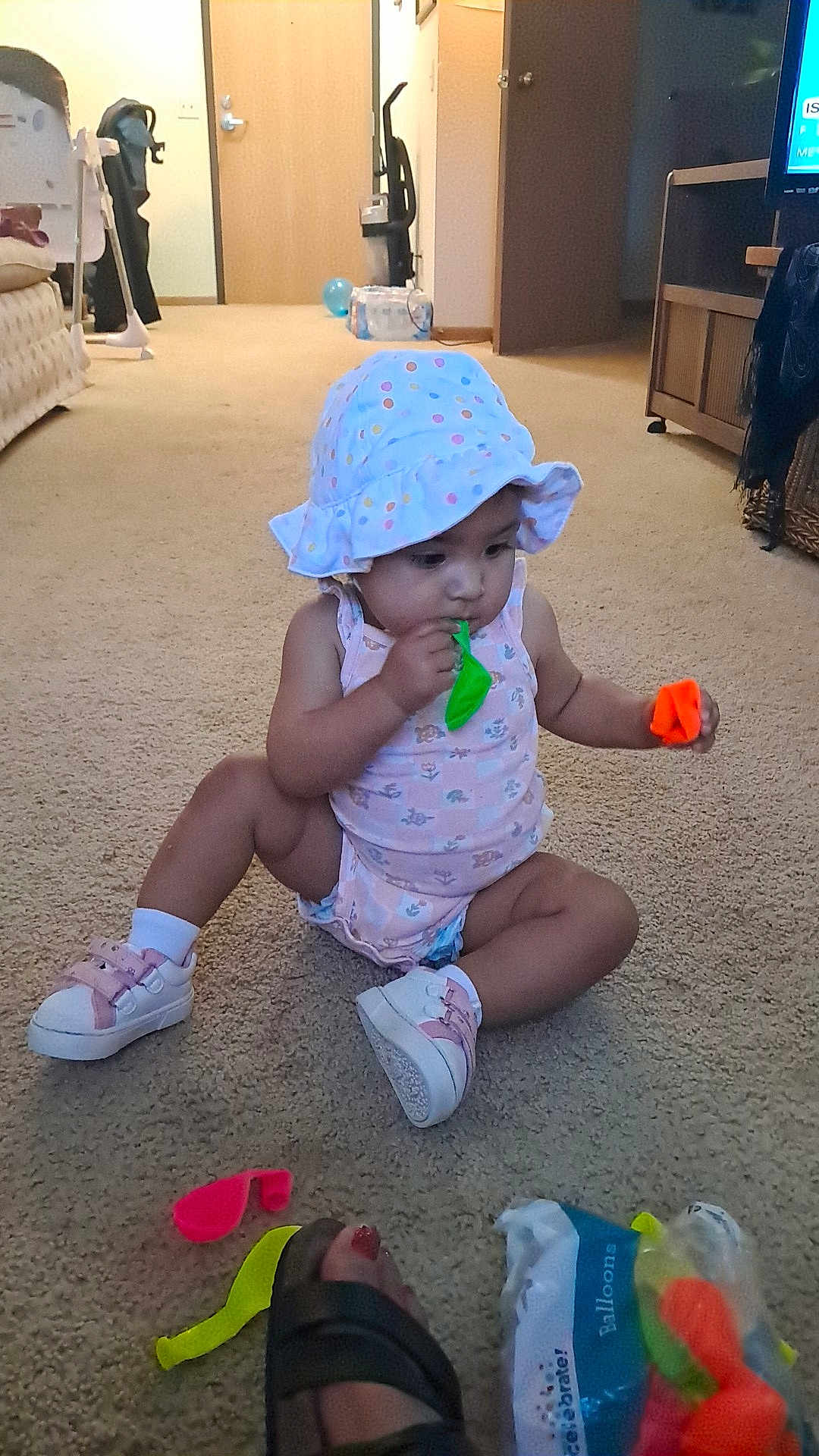 Ivy'Ann is registered to the contest to win money with this photo: toddler, child, balloon, hat, carpet, indoor, footwear, toy, sandal, pink, polka_dot, sitting, curious, home, living_room, door, plastic_bag, colorful, floor, person