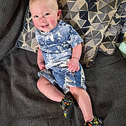 Benjamin is registered to the contest to win money with this photo: toddler, child, smile, sitting, couch, pillow, patterned_pillow, blue_clothing, shorts, shirt, footwear, colorful_shoes, indoor, happy, rosy_cheeks, baby, person, portrait, cute, casual_clothing