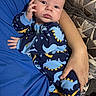 Benjamin is registered to the contest to win money with this photo: baby, infant, dinosaur_onesie, blue_clothing, hand, face, adult_arm, couch, fabric_pattern, curious_expression, cradling, indoor, person, child, snug, cozy, portrait, newborn, cute, soft_lighting