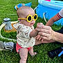 baby, child, sunglasses, yellow_sunglasses, grass, hand, adult_hand, sitting, outdoor, inflatable_pool, toy, shoe, person, shorts, hoodie, summer, playtime, sunny, fun, park