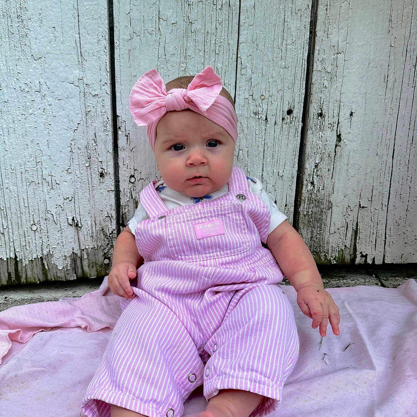Aylah Mae is registered to the contest to win money with this photo: baby, barefoot, casual, child, closeup, curious_expression, cute, headband, infant, natural_light, outdoor, overalls, person, pink_blanket, pink_bow, portrait, sitting, soft_texture, striped_clothing, wooden_wall