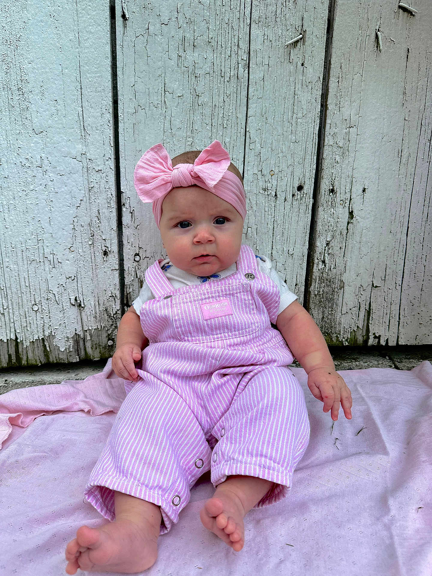 Aylah Mae is registered to the contest to win money with this photo: baby, pink_bow, headband, overalls, striped_clothing, pink_blanket, wooden_wall, outdoor, curious_expression, barefoot, child, person, infant, sitting, soft_texture, closeup, cute, casual, natural_light, portrait