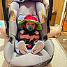 baby, car_seat, pacifier, hat, infant, child, adult, smiling, indoor, wood_paneling, person, footwear, sneakers, fabric, seat, handle, floor, mattress, home, cute