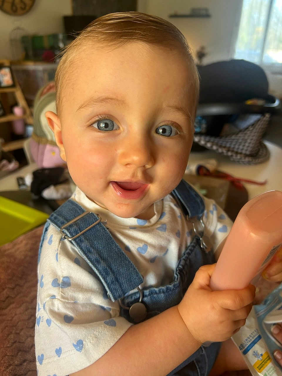 Alba participe au concours pour gagner de l'argent avec cette photo : baby, child, blue_eyes, denim_overalls, heart_pattern, smiling, indoor, holding, bottle, cute, portrait, happy, toddler, short_hair, skin, hand, clothing, face, person, home